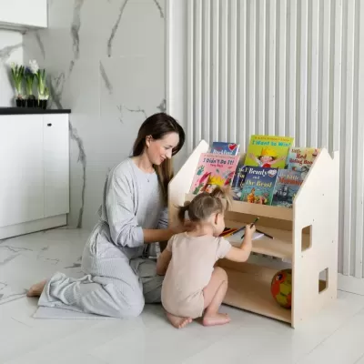 Estante infantil de madeira clara com livros e bola, mulher e criança desenham no chão em ambiente interior