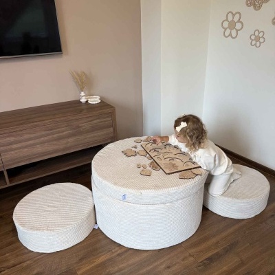 Mesa redonda e pufes bege em sala com chão de madeira e parede decorada, criança brinca com jogo de peças de madeira