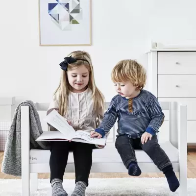 Menina e menino sentados num berço cama branco a ler um livro num quarto com chão de madeira.
