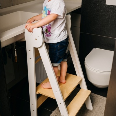 Criança de pé num banco-escalada branco com degraus de madeira clara junto a lavatório e sanita branco