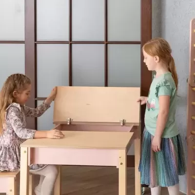Mesa e banco infantil em madeira clara e rosa com tampo articulado e duas crianças num interior com porta de vidro e parede cinza