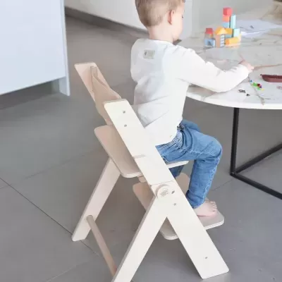 Criança sentada numa cadeira infantil de madeira clara junto a uma mesa de mármore com brinquedos coloridos