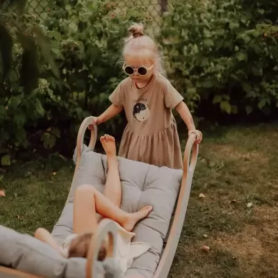 Duas crianças a brincar numa cadeira balanço de madeira com almofadas cinzentas num jardim