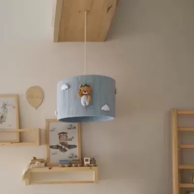 Candeeiro de teto azul claro com ursinho de peluche e nuvens, em quarto infantil com prateleiras de madeira e quadros.