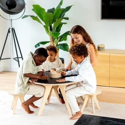Crianças e mulher sentados em volta de mesa pequena com base de madeira clara num ambiente interior moderno