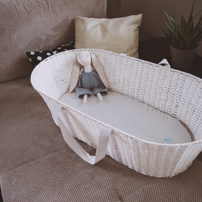 Cesta de bebé branca com colchão cinza e boneca de pano sobre sofá
