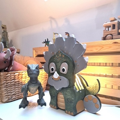 Dois brinquedos de dinossauros, um verde estilizado e outro cinzento realista, sobre mesa de madeira com outros brinquedos ao fundo