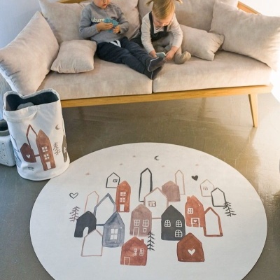 Tapete redondo branco com casas coloridas em chão cinza junto a sofá bege e duas crianças sentadas