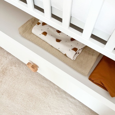 Roupas de bebé dobradas numa gaveta branca sob um berço branco e um tapete peludo bege no chão.