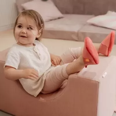 Criança sorridente sentada num sofá rosa claro com roupa clara e meias cor coral