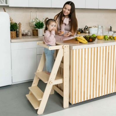 Criança em cadeirão de madeira e mulher na cozinha com frutas e sumo no balcão.