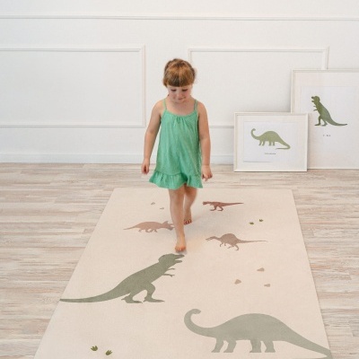 Tapete infantil bege com dinossauros verdes e castanhos, criança de vestido verde em cima e molduras com imagens de dinossauros ao fundo.