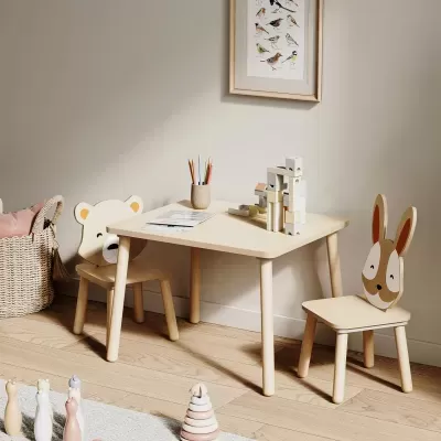 Mesa e cadeiras infantis em madeira clara com encostos decorados, lápis coloridos e blocos de madeira na mesa, ambiente com chão e parede claras.