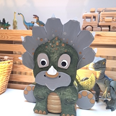 Figura brinquedo dinossauro verde em madeira com olhos grandes, sobre mesa branca com outros brinquedos ao fundo