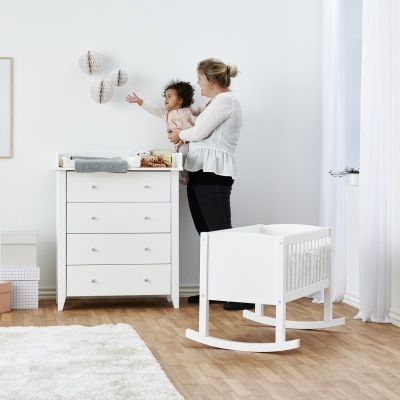 Cômoda branca de madeira com gavetas e berço branco tipo baloiço num quarto de bebé com chão madeira e parede branca