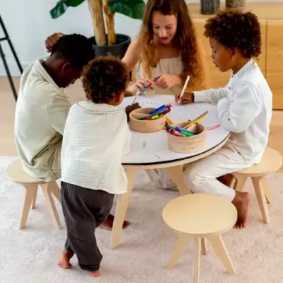 Crianças a desenhar numa mesa redonda com bancos de madeira clara