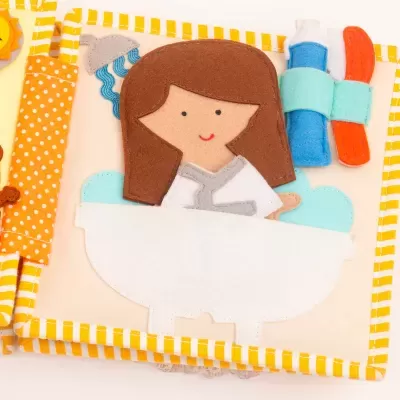 Livro de pano infantil com menina numa banheira e toalhas coloridas