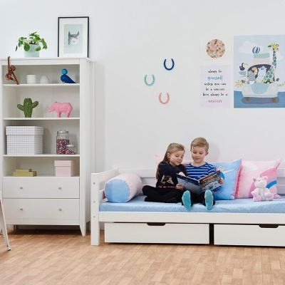 Quarto infantil com cama branca, almofadas coloridas, estante com brinquedos e duas crianças a lerem.