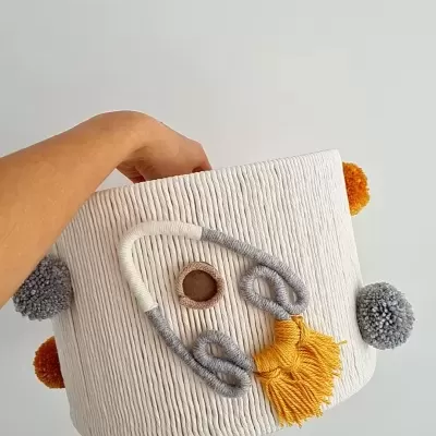 Cesto de tecido branco com detalhes em cordão cinza e amarelo e pompons coloridos