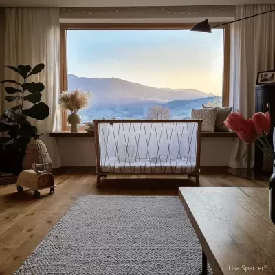 Berço de bebé em madeira clara próximo a janela grande com vista para montanhas ao pôr do sol num quarto acolhedor.