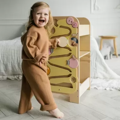 Brinquedo de atividade em madeira com painel amarelo e elementos rosa, em quarto claro com criança brincando