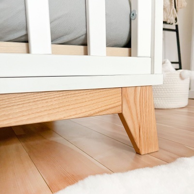 Detalhe da base de um berço branco com pernas de madeira clara sobre chão de madeira clara