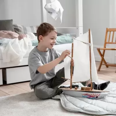 Menino sentado no chão a brincar com barco de vela em madeira num quarto com cama e cadeira de madeira.