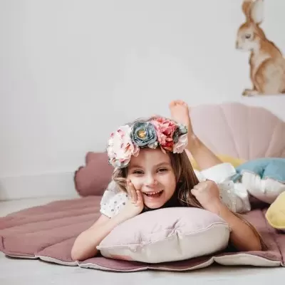 Criança sorridente deitada sobre tapete com almofada e coroa de flores, parede branca com adesivo de coelho