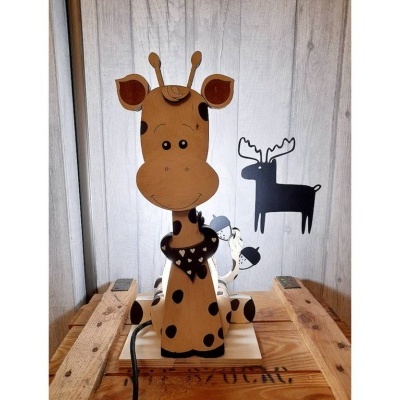 Abajur infantil girafa madeira com manchas castanho claro e escuro, cachecol preto, olhos fechados, em palete de madeira com parede branca e decalques de cervos preto