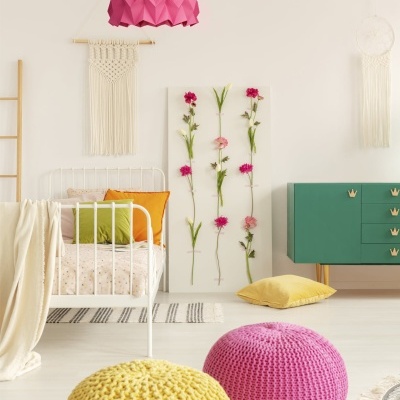 Quarto decorado com cama branca, pufes tricotados, candeeiro rosa e decoração floral