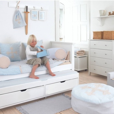 Quarto infantil com cama branca, almofadas azuis, cadeira e pufe e cómoda branca com cestos.