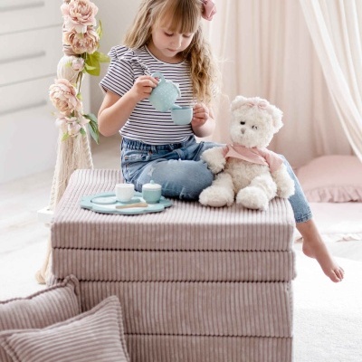 Criança brinca com chávena azul em colcha rosa riscada com almofada e peluche ao lado