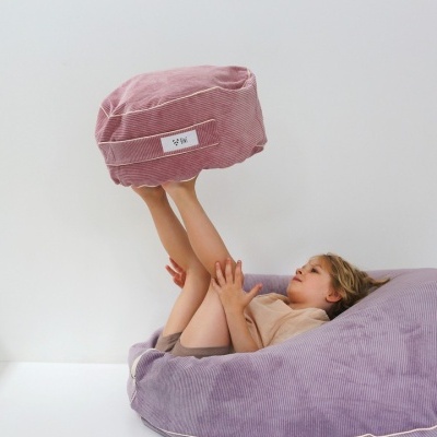 Criança deitada num puff lilás grande segurando um puff rosado redondo com etiqueta 'VIKI'