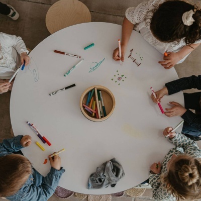 Mesa redonda branca com crianças a desenhar com canetas e pincéis coloridos