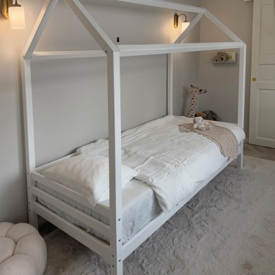 Cama infantil branca em forma de casa com roupa branca e manta bege em quarto neutro