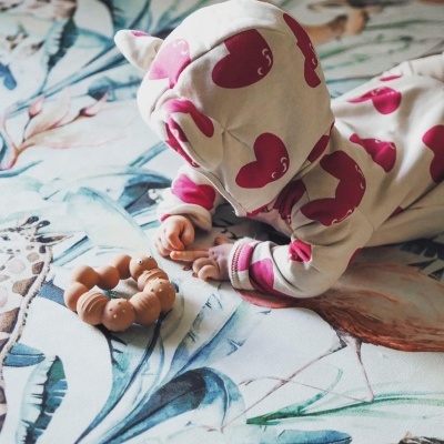 Bebé de pijama com corações rosa e capuz com orelhas deitado em tapete floral com brinquedo silicone