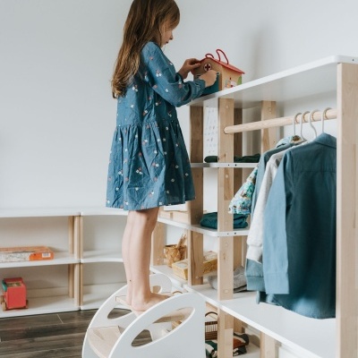 Menina de vestido azul brinca com casinha de madeira vermelha numa estante de madeira clara num quarto com chão de madeira escura