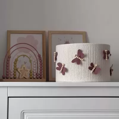 Abajur branco com borboletas cor-de-rosa e pérolas numa cómoda branca com quadros coloridos atrás.