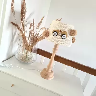 Abajur de madeira e tecido com pompons e botões sobre cómoda branca, ao lado de vaso de vidro com ramos secos.