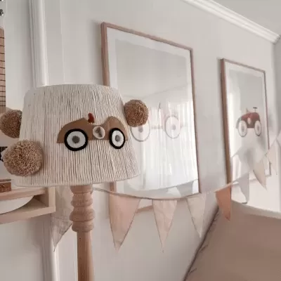 Abajur de madeira com cúpula bege e aplique de carrinho junto a quadros e bandeirolas numa sala clara.