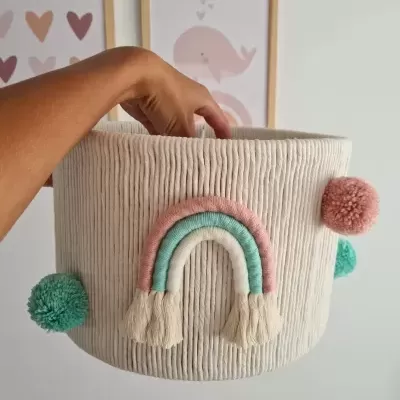 Cesto de tecido cru com arco-íris colorido e pompons decorativos suspenso por uma mão