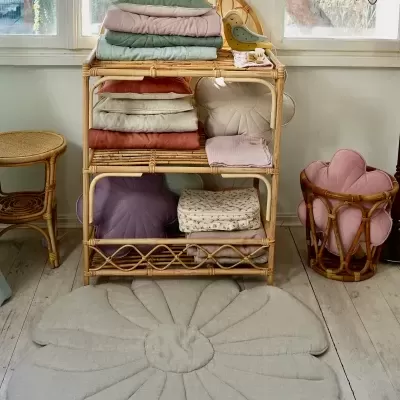 Estante de vime com almofadas e mantas coloridas, tapete floral cinza no chão e decoração de flores na janela