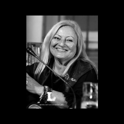 Mulher sorridente loira com blusa escura e pulseiras sentada atrás de microfone