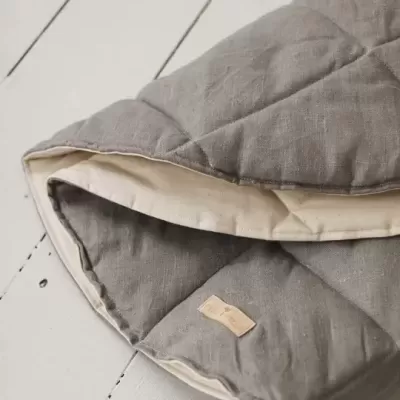 Colcha cinzenta matelassada com acabamento cru sobre piso branco.