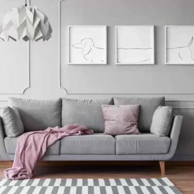 Sofá cinzento claro com almofadas e manta rosa numa sala com moldura na parede e quadros minimalistas