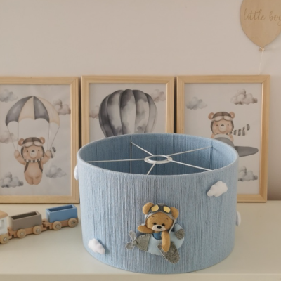 Abajur de teto azul claro com urso aviador em avião e nuvens, em ambiente infantil com quadros e brinquedos em madeira