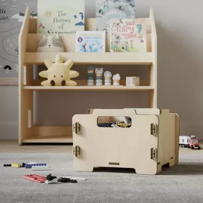 Caixa de armazenamento de brinquedos em madeira clara com tapete cinzento e estante de madeira com livros e brinquedos ao fundo.