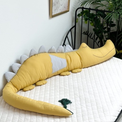 Almofada grande em forma de crocodilo amarela com detalhes cinza em cama branca