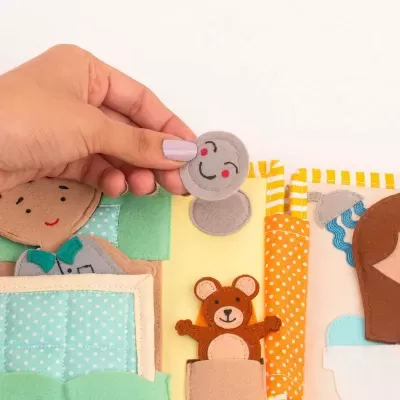 Livro têxtil infantil com bonecos de tecido, cama verde, urso castanho e carinha sorridente cinza