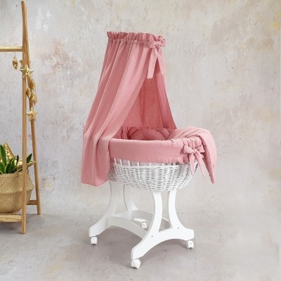 Berço oval branco com suporte branco e têxtil rosa com dossel num ambiente neutro com estante de madeira e plantas